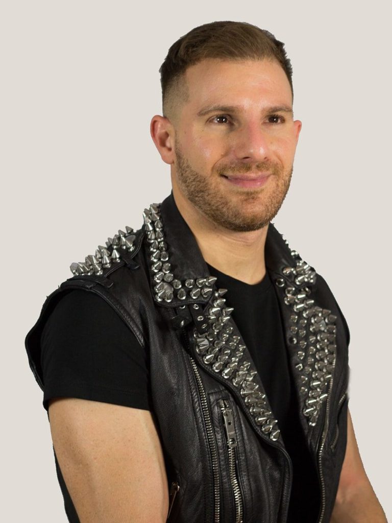 A photograph of Ben Barry taken by Akash Rai. He gazes off to the right hand side of the photograph and he has a slight smile on his face. He has short brown hair with a skin fade on the sides, dark brown eyes and white skin with a shortly trimmed brown beard. He wears a black leather vest that has shiny silver spikes on the shoulders, the collar and the lapel over a black t-shirt.